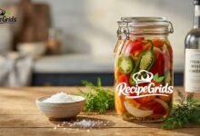 A sealed glass jar filled with colorful homemade pickled peppers, onions, and carrots sitting next to a bottle of premium white vinegar and coarse salt.