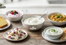 A rustic wooden table displaying various homemade dishes including plain yogurt, berry yogurt, creamy chicken curry, tzatziki with pita bread, frozen yogurt bark, and sliced lemon cake.
