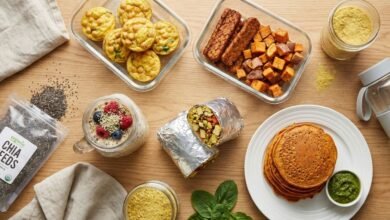 Flat lay photograph of high protein vegan breakfast meal prep on a wooden counter, showing tofu burritos, chickpea muffins, tempeh sweet potato hash, savory red lentil pancakes, and overnight oats without eggs.