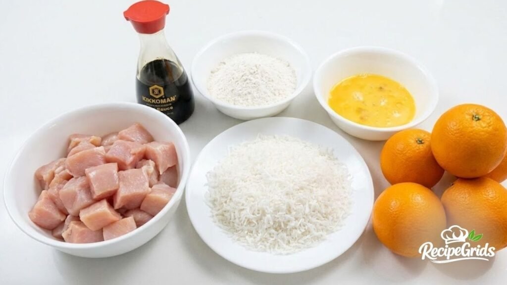 Ingredients needed to make coconut chicken that's like orange chicken, including raw diced chicken, shredded coconut, fresh oranges, flour, eggs, and soy sauce.