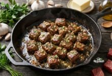 Sizzling cast iron skillet filled with golden-brown keto garlic butter steak bites, garnished with fresh parsley, surrounded by whole garlic bulbs, butter blocks, and olive oil on a rustic wooden table.