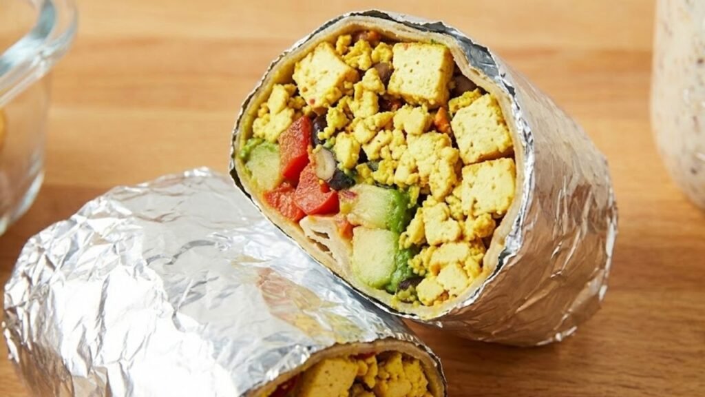 A breakfast burrito, cut in half and wrapped in aluminum foil, showing a filling of tofu scramble, black beans, diced red peppers, and avocado.