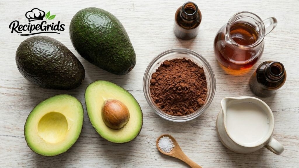 An overhead photograph on a light wood grain background showing the ingredients for a vegan avocado chocolate mousse recipe. Elements include whole and halved fresh avocados, a glass bowl of cocoa powder, a pitcher of maple syrup, a mug of plant milk, vanilla extract bottles, and a wooden spoon with sea salt.
