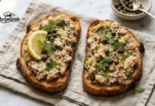 Two slices of toasted sourdough bread generously topped with chunky tuna and white bean smash, garnished with capers, fresh dill, olive oil, and a lemon wedge on a linen napkin.