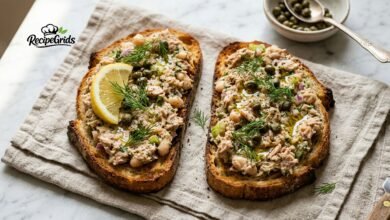 Two slices of toasted sourdough bread generously topped with chunky tuna and white bean smash, garnished with capers, fresh dill, olive oil, and a lemon wedge on a linen napkin.
