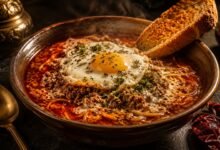 A steaming, dark red bowl of the best sopa criolla near me, featuring seasoned ground beef, fine angel hair noodles, and evaporated milk, topped with a perfectly fried egg and crispy toasted bread.